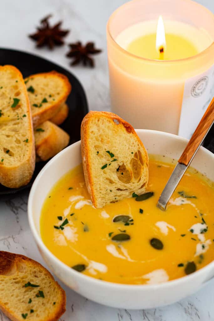 Creamy butternut squash bisque served in a white bowl, garnished with pumpkin seeds, fresh herbs, and a swirl of cream, with a slice of crusty bread resting on the side, candle and bread plate in the background.