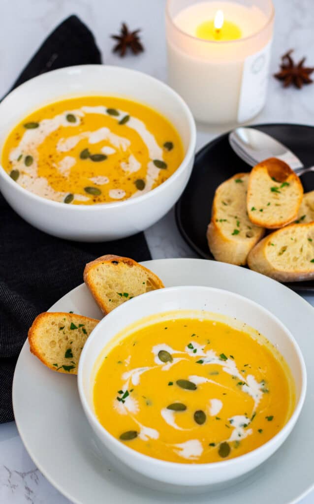 Two bowls of creamy butternut squash bisque garnished with pumpkin seeds, fresh herbs, and cream, served with slices of crusty bread on a fall-inspired table with a lit candle in the background.