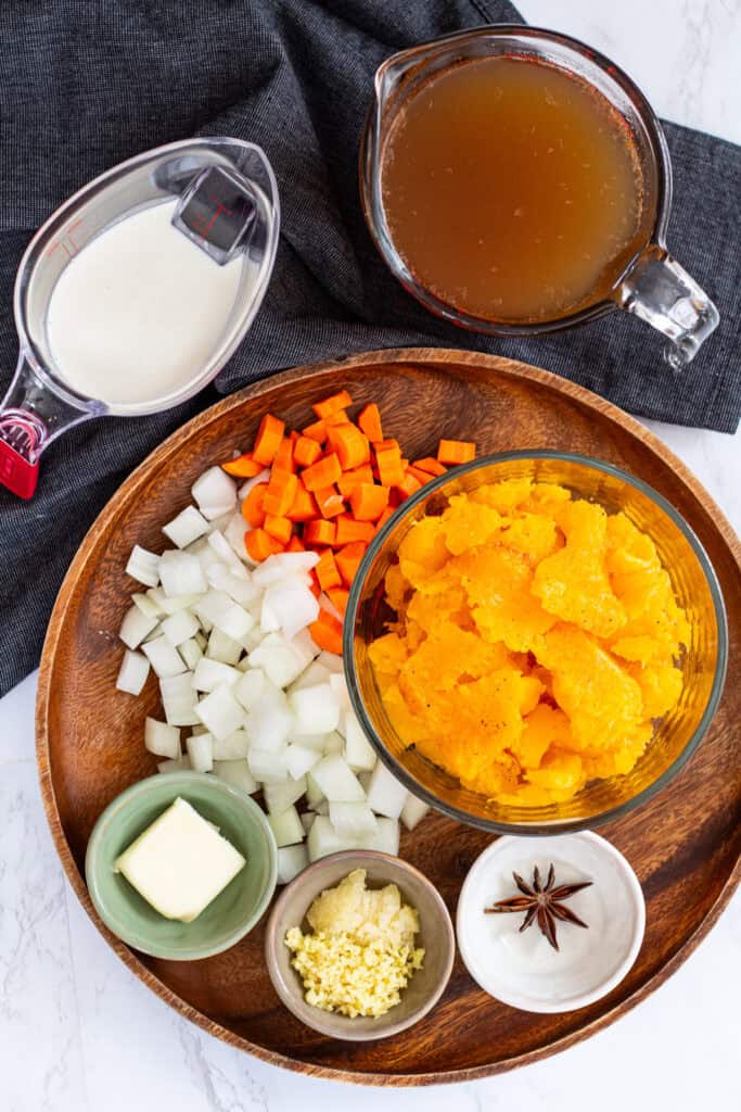 Overhead view of butternut squash bisque ingredients on a wooden platter, including roasted butternut squash, carrots, onions, butter, minced garlic and ginger, star anise, broth, and cream.