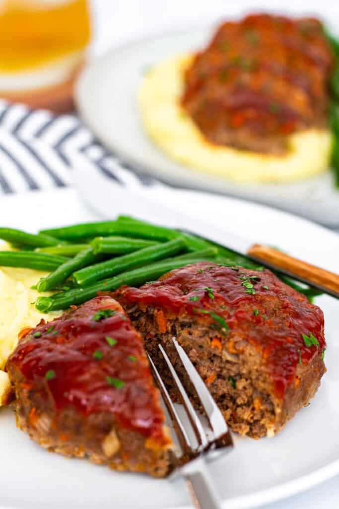 Whole glazed mini meatloaf cut open with a fork, served alongside mashed potatoes and green beans