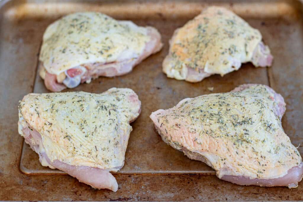 Raw chicken thighs on a sheet pan coated under the skin with dried herb butter for roasting.