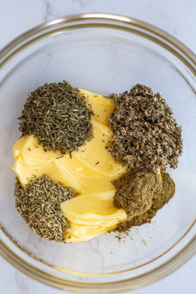Softened butter in a glass bowl with dried rosemary, thyme, and sage, ready to be mixed into dried herb butter for poultry.