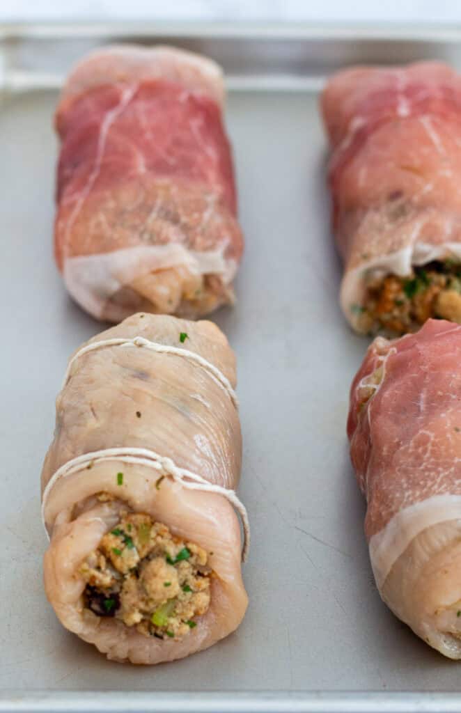 Chicken breasts rolled with stuffing, wrapped in prosciutto, or tied with kitchen twine on a baking sheet.