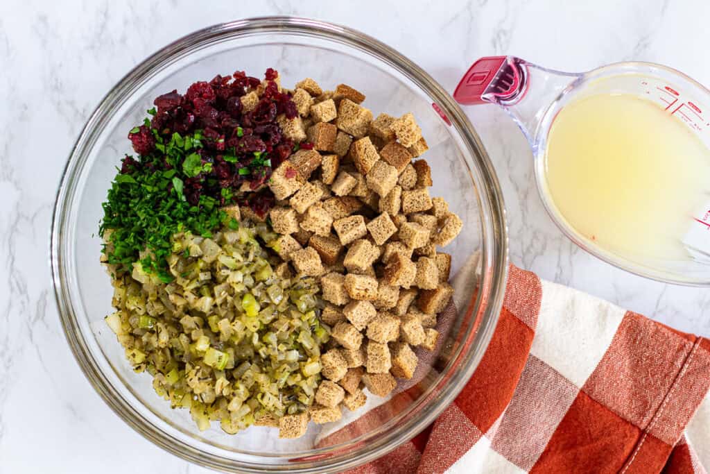 Bowl of cubed bread, celery, onion, parsley, and cranberries with broth on the side, ready to make homemade stuffing.