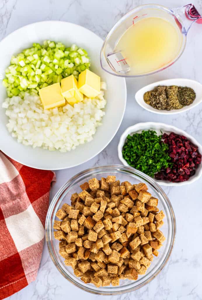 Flatlay of stuffing ingredients including bread cubes, celery, onion, butter, broth, herbs, parsley, and dried cranberries.