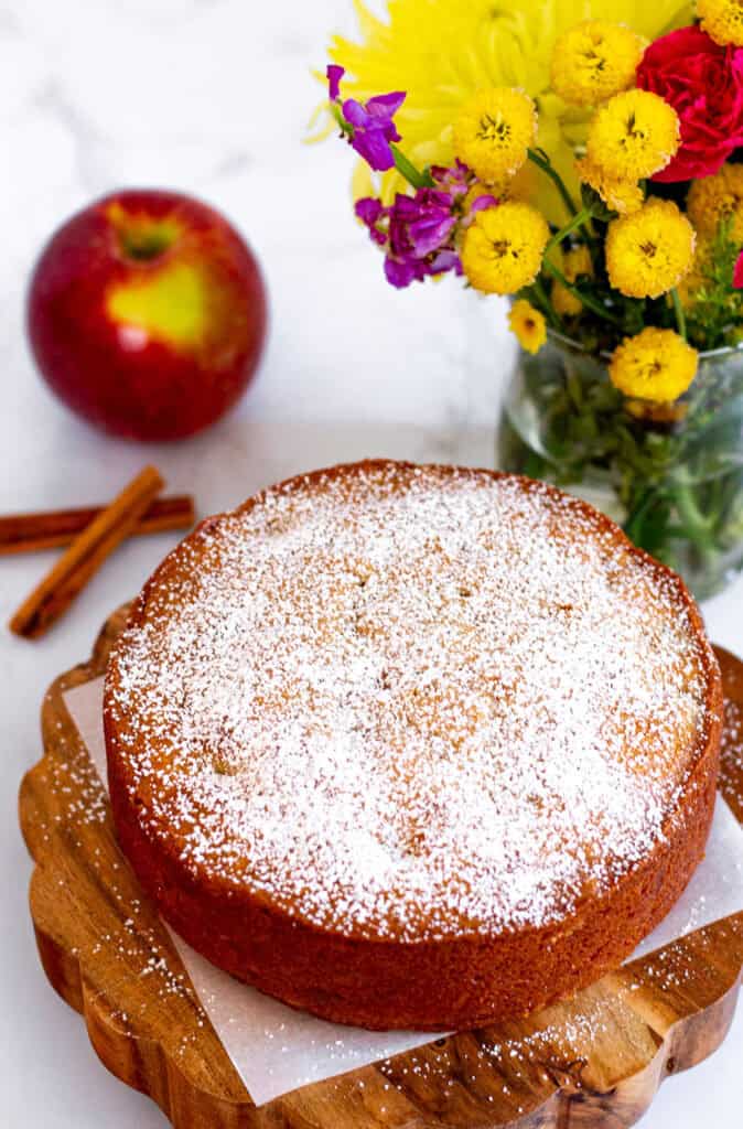 Warm spiced apple cake dusted with powdered sugar on a wooden cake stand with flowers and apples
