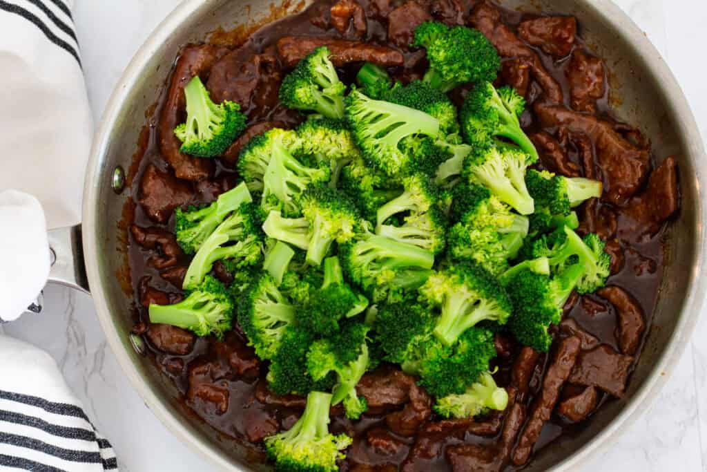 Blanched broccoli added on top of cooked beef in sauce, ready to toss together for beef and broccoli.