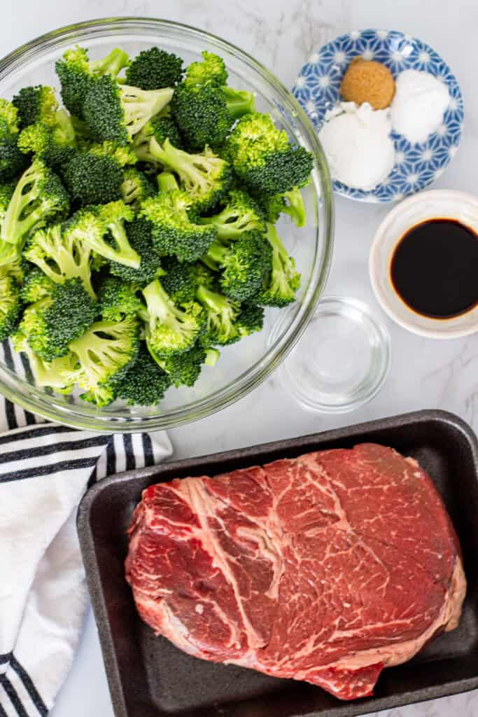 Overhead shot of broccoli florets, a cut of beef, soy sauce, brown sugar, cornstarch, and seasonings arranged on a marble surface for beef and broccoli recipe.