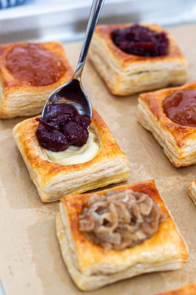 Spoon adding cranberry sauce filling to brie-filled puff pastry bites on a baking sheet.
