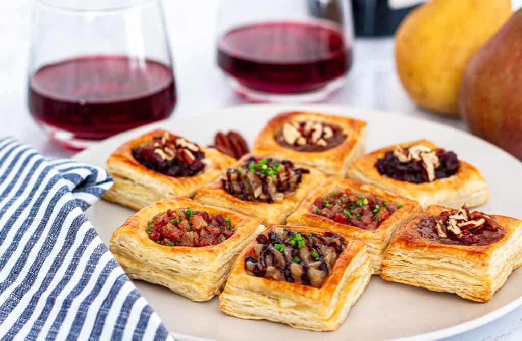 Close-up of puff pastry brie bites on a plate showing flaky layers and assorted sweet and savory toppings.