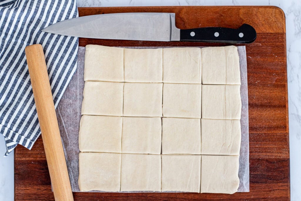 Puff pastry sheet cut into small squares on a wooden board with knife and rolling pin, ready for baking.