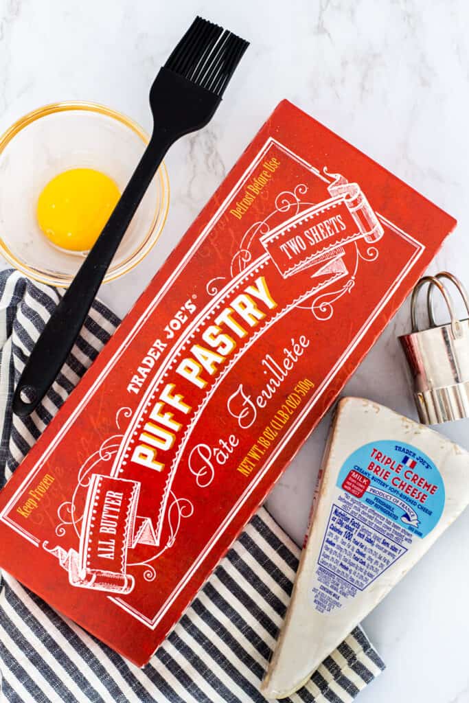Trader Joe’s puff pastry box with a wedge of brie, egg wash, and pastry brush on a striped napkin.