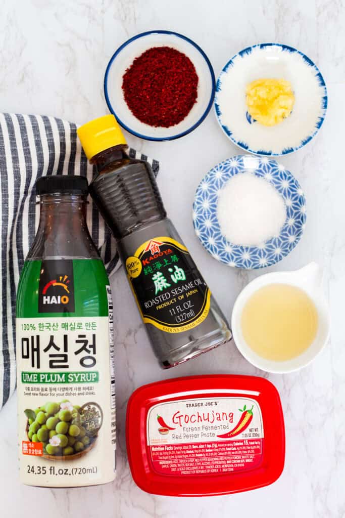 Ingredients for Korean cucumber salad including gochujang, sesame oil, maesil-aek, garlic, and vinegar arranged on a marble surface.
