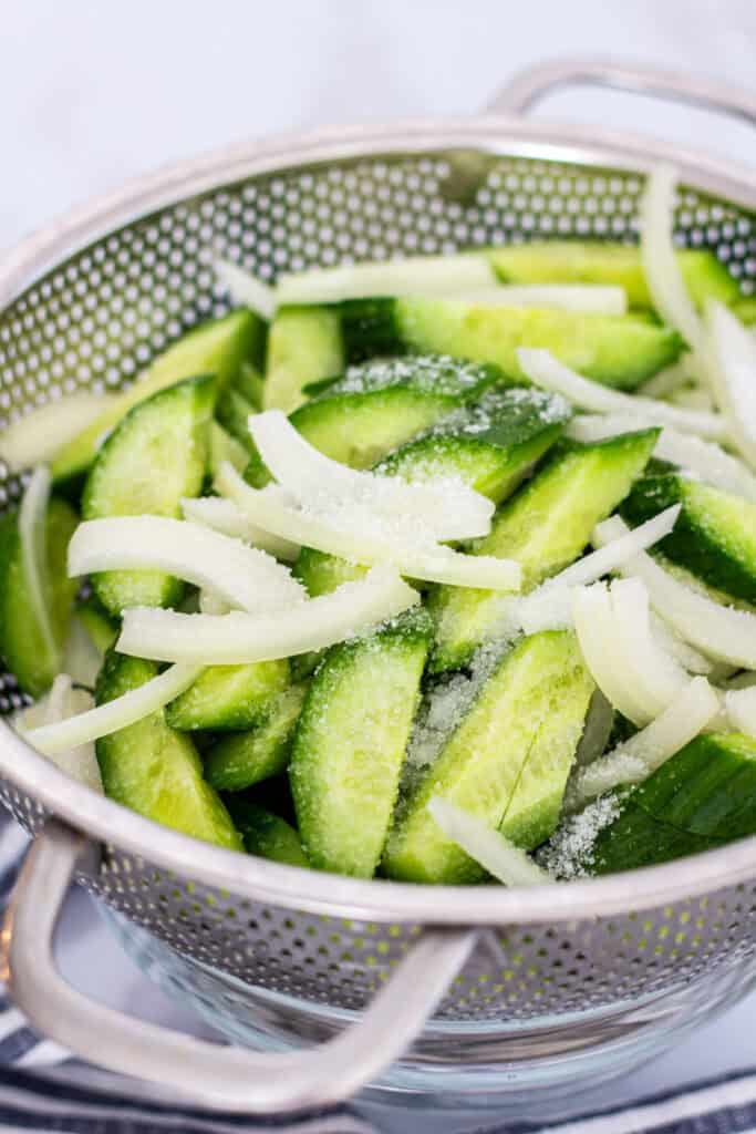 Sliced cucumbers and onions sprinkled with salt in a colander to draw out moisture for Korean cucumber salad.