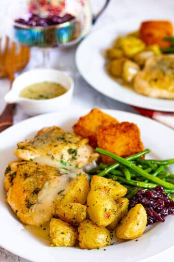 Close-up of a small Thanksgiving dinner plate with chicken thighs, mashed potatoes, green beans, and cranberry sauce topped with gravy.
