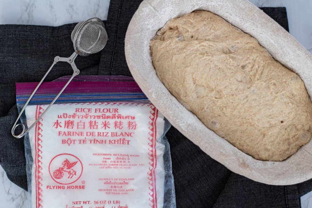 Shaped sourdough discard dough resting in a rice-floured banneton with rice flour and a sifter beside it.
