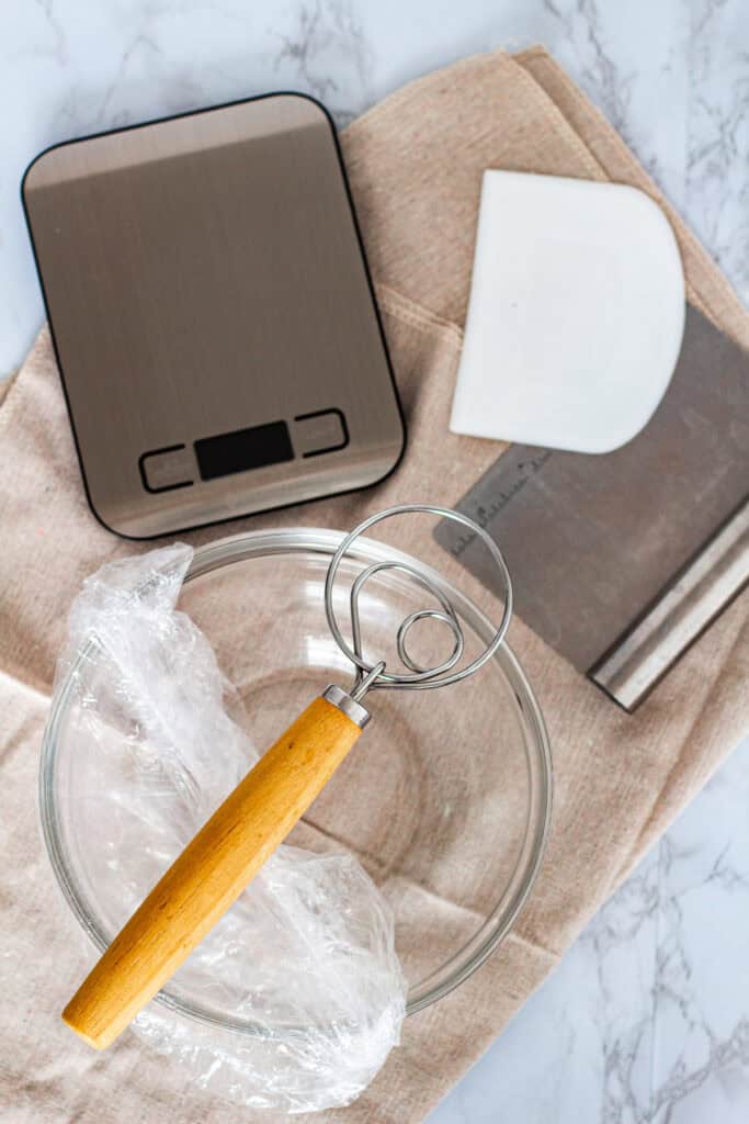 Mixing tools for making sourdough discard bread, including a digital scale, dough whisk, bowl scraper, and glass bowl.