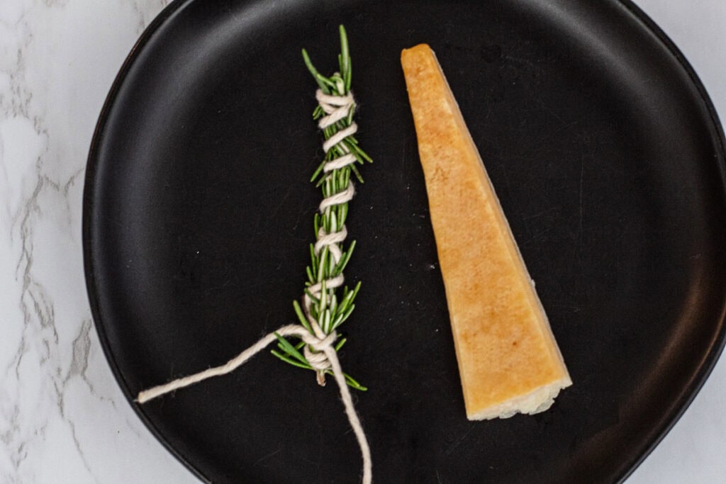 Rosemary tied with kitchen twine next to a parmesan rind on a black plate.