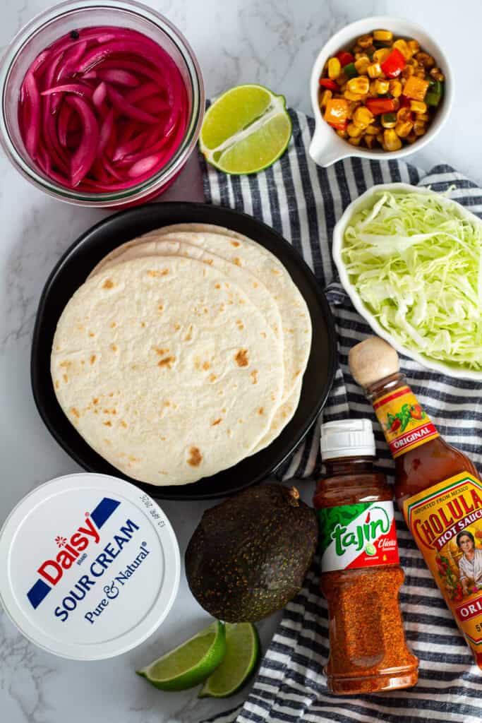 Ingredients for blackened fish tacos including tortillas, shredded cabbage, pickled red onions, corn, avocado, and seasonings.