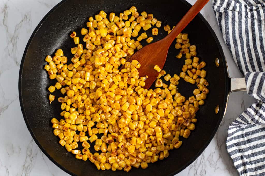 overhead view of sauteed corn cooking in a skillet with a wooden spoon