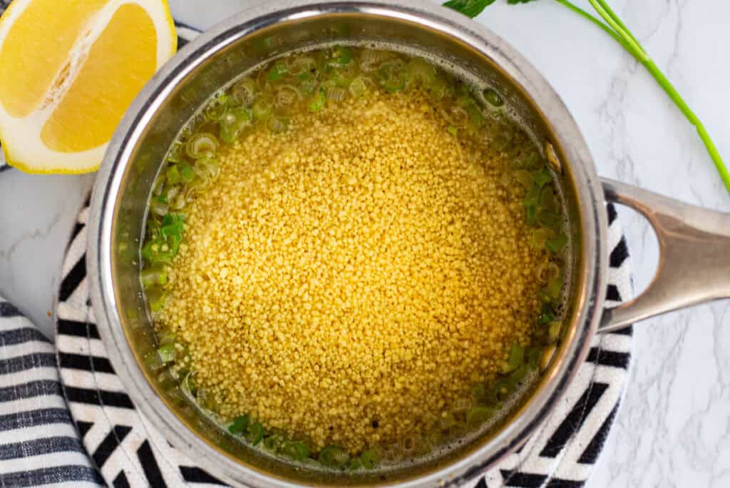 Couscous soaking in warm broth with sliced green onions in a small pot