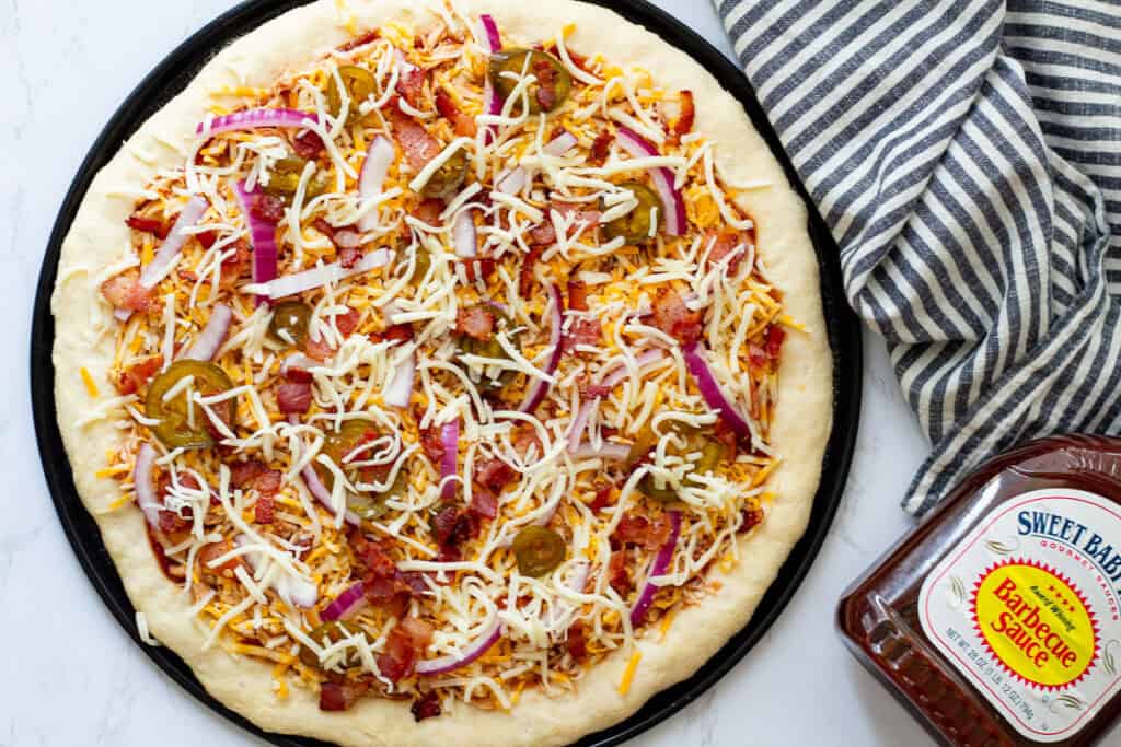 BBQ chicken and bacon pizza topped with cheese, bacon, red onion, and pickled jalapeños before baking.