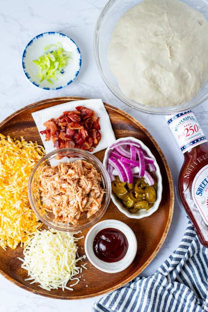 Ingredients for making BBQ chicken and bacon pizza, including pizza dough, shredded chicken, bacon, cheese, red onion, pickled jalapeños, green onions, BBQ sauce, and mozzarella.