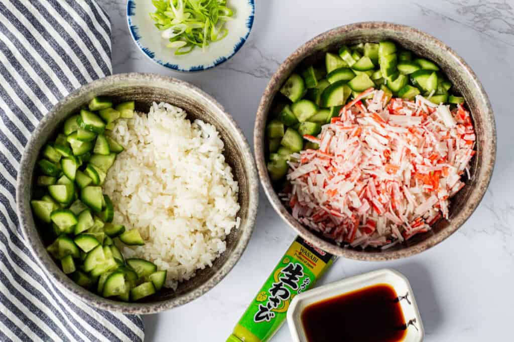Sushi rice topped with diced cucumber and imitation crab while assembling a California Roll Bowl.
