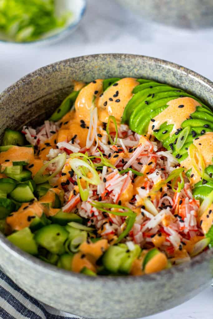 Close-up of a California Roll Bowl topped with imitation crab, avocado, cucumber, spicy mayo, green onion, and black sesame seeds.