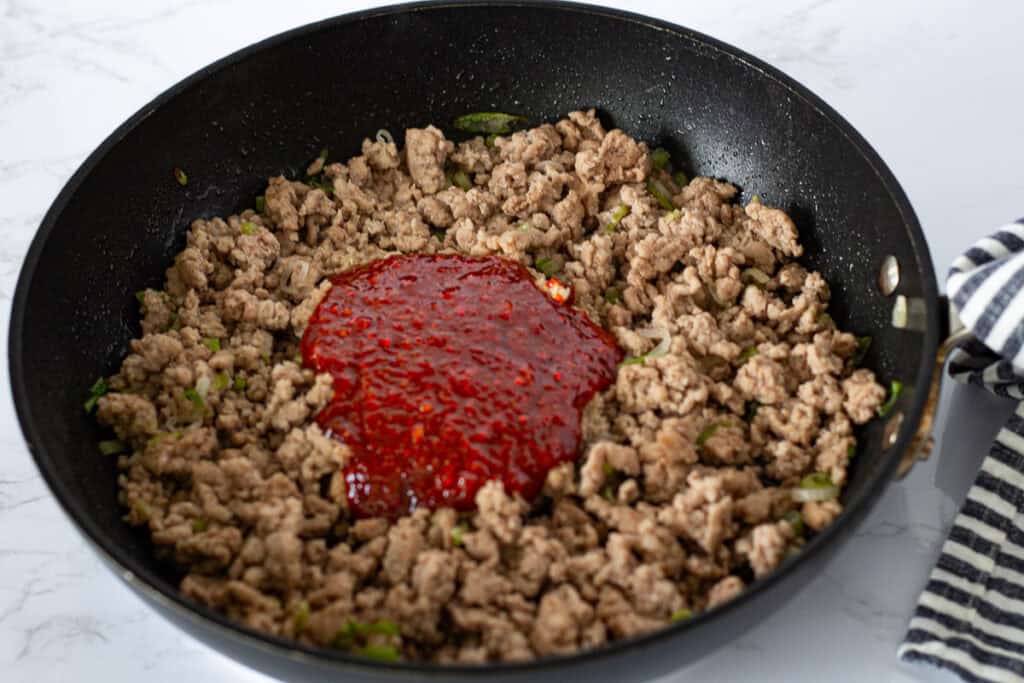 Pouring the gochujang sauce over cooked ground pork in a skillet.