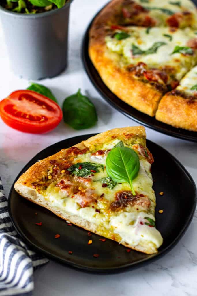 Slice of pesto tomato pizza topped with melted mozzarella, fresh basil, Parmesan, and red pepper flakes.