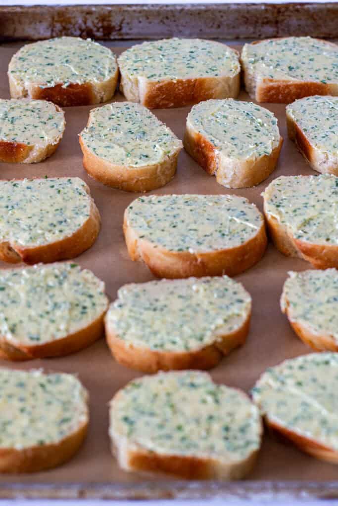 Garlic butter spread to the edges of French bread slices arranged on a parchment-lined baking sheet