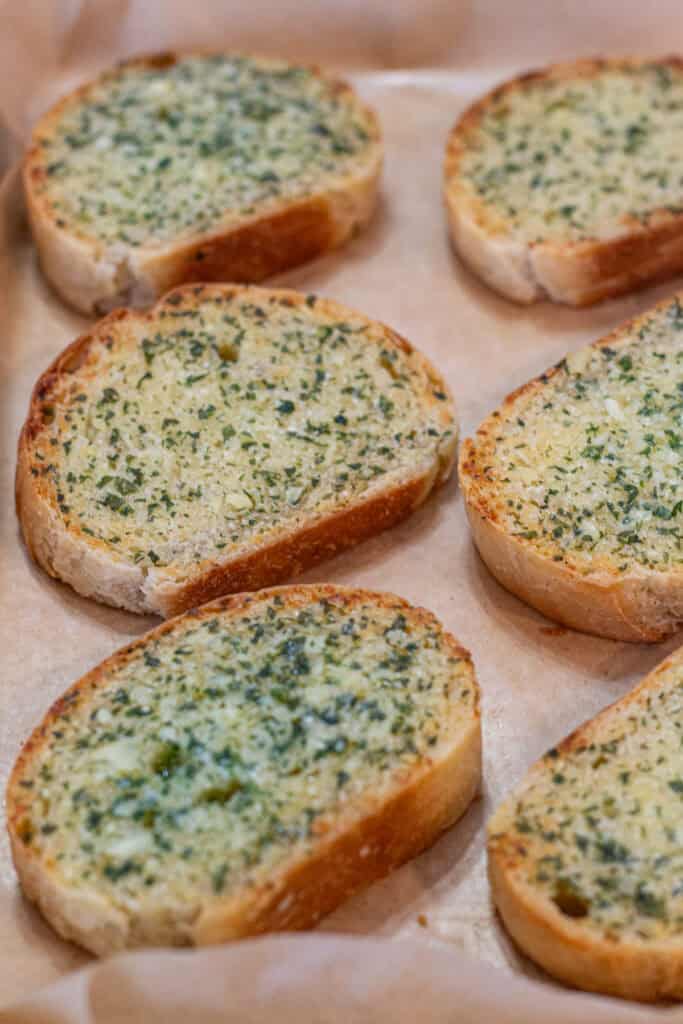 Six slices of homemade garlic bread with golden edges, garlic butter, and parsley on a parchment-lined baking sheet.
