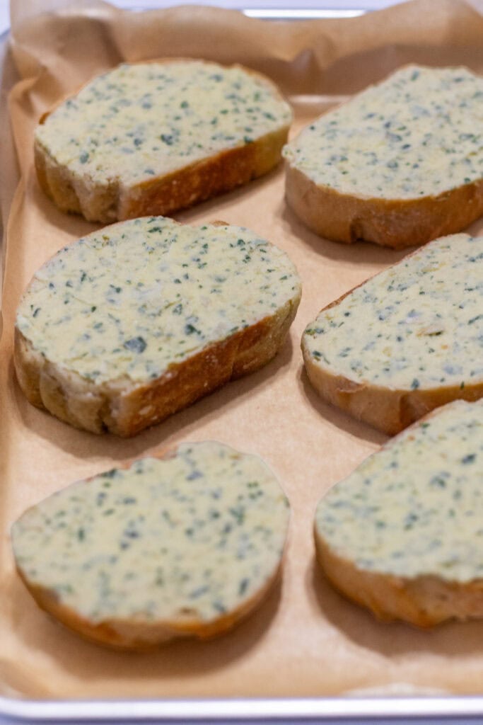 Frozen garlic bread slices topped with garlic-Parmesan butter and parsley on a parchment-lined baking sheet, ready to bake straight from the freezer.