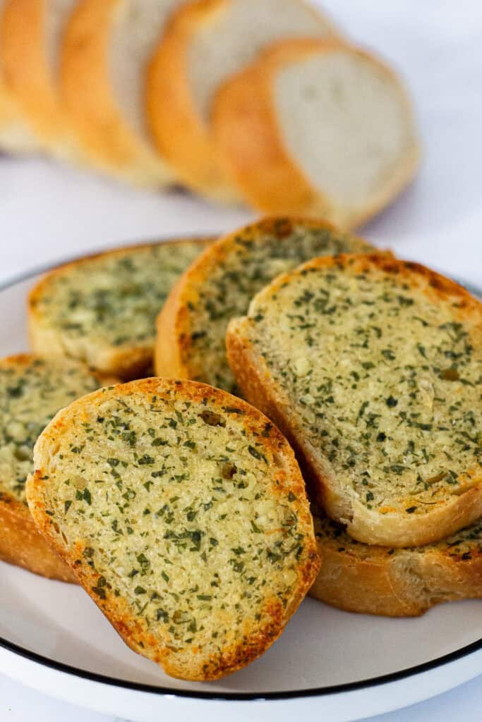 Golden, toasted garlic bread slices arranged on a white plate with a loaf of French bread in the background.