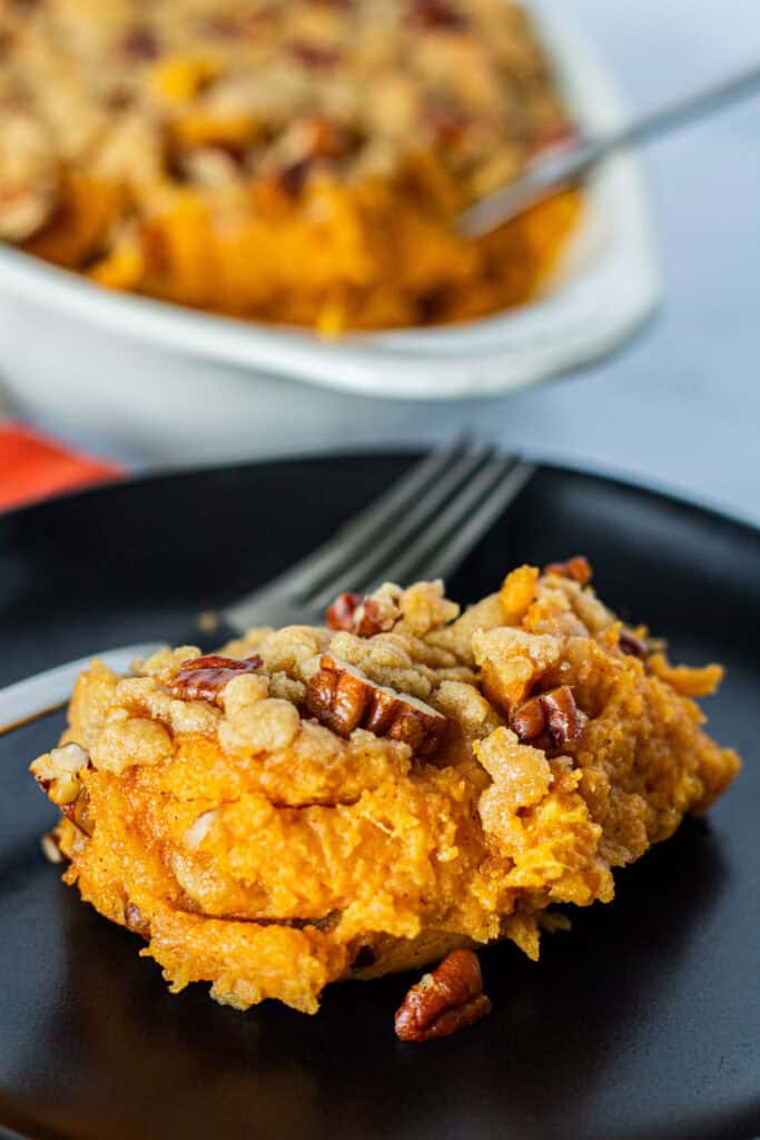 A serving of sweet potato casserole with brown sugar pecan crumble on a black plate, with the baking dish in the background.