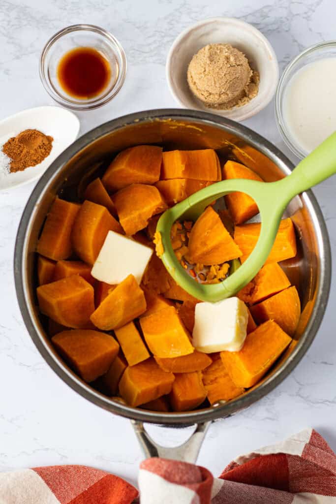 Cooked sweet potato chunks in a saucepan with butter and a potato masher, surrounded by filling ingredients.
