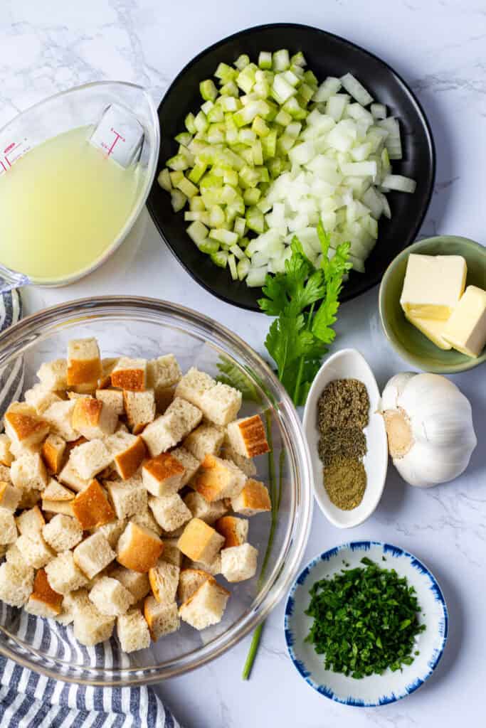 Bread cubes, diced onion and celery, butter, chicken broth, dried herbs, garlic, and fresh parsley arranged for homemade stuffing.