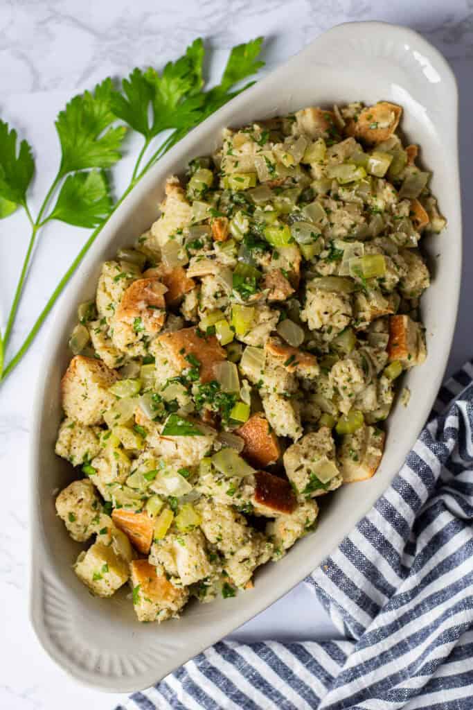 Bread cubes mixed with sautéed onion, celery, herbs, and parsley in a small white oval baking dish, ready for the oven.