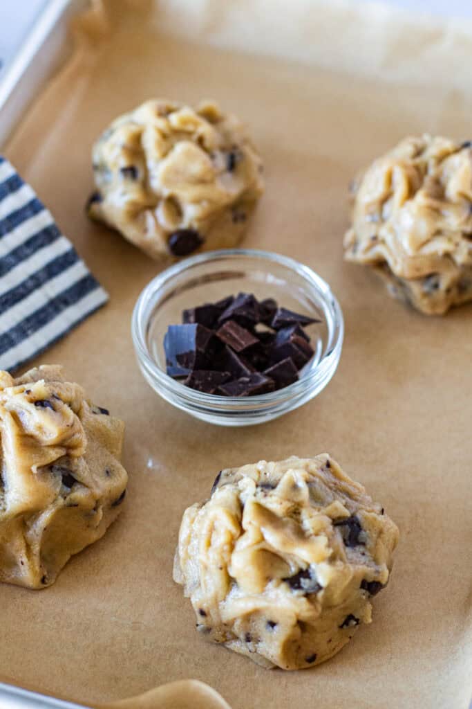 Four roughly shaped chocolate chip cookie dough mounds on a parchment-lined baking sheet before chilling